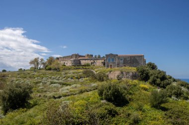 Çok uzun bir geçmişi olan, yeşilliklerle kaplı bir tepede inşa edilmiş bir Milazzo kalesi. Arka planda Tyrhenian (Akdeniz) denizi. Beyaz bulutlu mavi gökyüzü. Milazzo, Sicilya, İtalya.