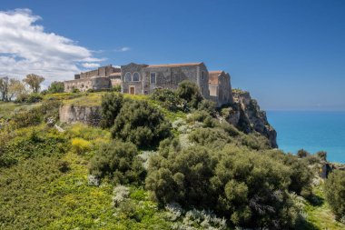 Çok uzun bir geçmişi olan, yeşilliklerle kaplı bir tepede inşa edilmiş bir Milazzo kalesi. Arka planda Tyrhenian (Akdeniz) denizi. Beyaz bulutlu mavi gökyüzü. Milazzo, Sicilya, İtalya.