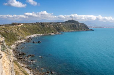 Tyrhenian (Akdeniz) denizi ve kıyı kayalarının durgun sularının parlak koyu turkuaz rengi. Beyaz bulutlu mavi gökyüzü. Milazzo, Sicilya, İtalya.