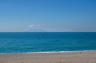 Çakıl taşlı kumsal. Tiren (Akdeniz) denizinin sakin turkuaz suyu. Ufuktaki Aeolian adaları. Küçük beyaz bulutlu mavi gökyüzü. Milazzo, Sicilya, İtalya.