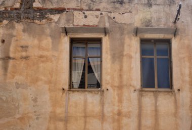 Eski, yıpranmış turuncu bir ev cephesi, kısmen çökmüş bir cephe. İki kapalı pencere, birinin perdesi var. Cefalu Caddesi, Sicilya, İtalya.