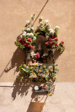 Çapraz gölge çizgisi olan bir evin aydınlanmış duvarı. Etrafta heykeli ve yapay çiçekleri olan bir tapınak. Cefalu, Sicilya, İtalya.