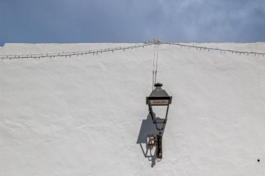 White wall with a traditional lantern. Cable with small lights. Everything with its shadow. Blue sky with white clouds. Teguise, Lanzarote, Canary Islands, Spain. clipart