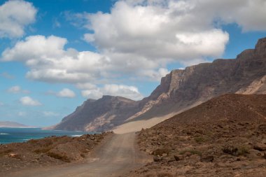 Atlantik Okyanusu kıyısındaki volkanik kayalar. Beyaz kumlu bir sahil. Kışın bulutlu bir gökyüzü. Famara, Lanzarote, Kanarya Adaları, İspanya.