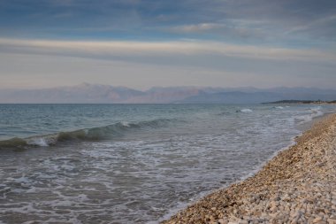 İyonik denizin küçük dalgaları. Çakıl taşlı plaj. Arkadaki Arnavut dağları. Işık bulutlu gökyüzü. Acharavi, Korfu, Yunanistan.