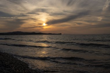 Small waves of the Ionic sea. Beach with pebbles. Mountain in the background. Sunset and cloudy sky. Reflection on the sea. Acharavi, Korfu, Greece. clipart