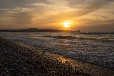 İyonik denizin küçük dalgaları. Çakıl taşlı plaj. Arka plandaki dağ. Gün batımı ve bulutlu gökyüzü. Denize yansıyor. Acharavi, Korfu, Yunanistan.