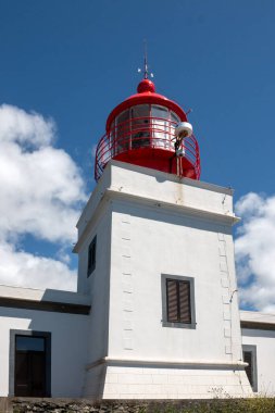 Beyaz yüzlü ve kırmızı kulesi olan bir deniz feneri inşa etmek. Bir uçurumun tepesinde. Açık beyaz bulutlu mavi gökyüzü. Ponta do Pargo, Madeira, Portekiz.