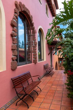 Wall of a building with arched windows. Empty wooden benches. Orange floor. Leaves of the plants in a botanical garden, Canico, Madeira, Portugal. clipart