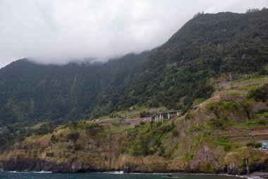 Volkanik kayalar ve kayalıklar taze yeşilliklerle kaplı. İlkbaharda bulutlu bir gün, dağın tepesinde bir bulutun içinde. Yol çizgisi. Seixal, Madeira, Portekiz.