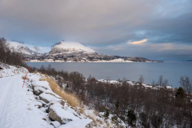 Snow-covered landscape. Mountains and a water of a Kvaerfjord. Colorful cloudy sky during sunrise/sunset. Harstad, Norway. clipart