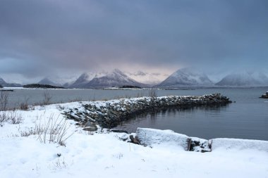 Snow-covered landscape. Mountains and a water of a Kvaerfjord. Colorful cloudy sky during sunrise/sunset. Harstad, Norway. clipart