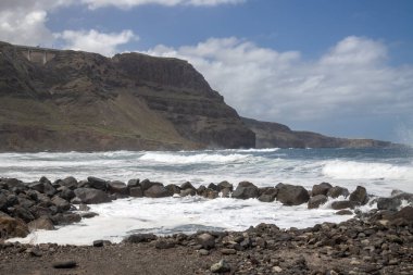 Atlantik Okyanusu dalgaları. Rocky Coast küçük bir havuz oluşturuyor. Taşlı plaj. Arka plandaki dağ. Beyaz bulutlu mavi gökyüzü. Gula, Las Palmas, Gran Canaria, Kanarya Adaları, İspanya.