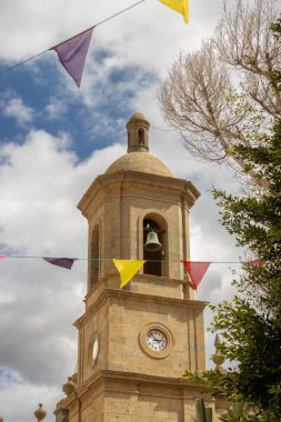 Aziz Sebastia Kilisesi (Iglesia de San Sebastian), çan kulesi ve saati ile. Mavi gökyüzü ve beyaz bulutlar. Renkli bayraklar. Aguimes, Las Palmas, Gran Canaria, Kanarya Adaları, İspanya.