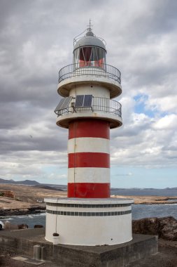 Deniz feneri (Faro de Arinaga) kırmızı ve beyaz çizgili, geleneksel tarzda. Beyaz bulutlu mavi gökyüzü. Atlantik Okyanusu kıyısında. Arinaga, Las Palmas, Gran Canaria, Kanarya Adaları, İspanya.