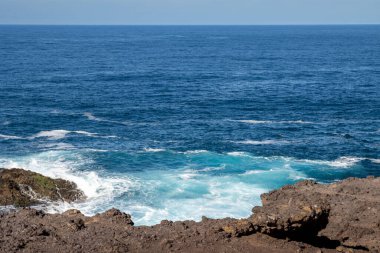 Karanlık volkanik kıyı kayaları. Atlas Okyanusu 'nu sakinleştir. Kayaların üzerinde dalgalar var. Mavi gökyüzü. Galdar, Las Palmas, Gran Kanarya, Kanarya Adaları, İspanya.