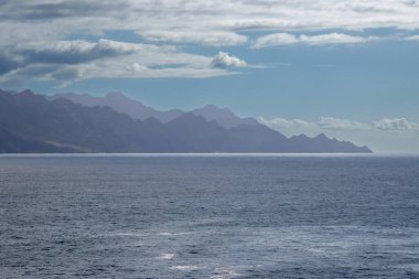 Sis içindeki karanlık volkanik kıyı dağları. Sakin Atlantik Okyanusu. Beyaz bulutlu mavi gökyüzü. Galdar, Las Palmas, Gran Kanarya, Kanarya Adaları, İspanya.