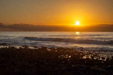 Atlantik Okyanusu sahilinde dalgalı volkanik çakıl taşları. Gün batımında bulutlu turuncu gökyüzü. Puerto de las Nieves, Las Palmas, Gran Canaria, Kanarya Adaları, İspanya.