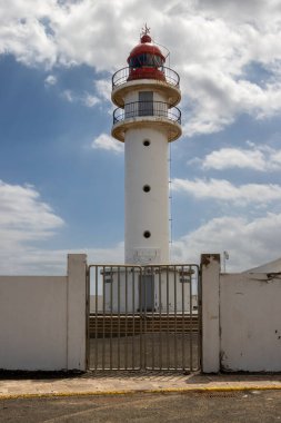 Beyaz yüzlü, kırmızı bluzlu, geleneksel tarzda bir deniz feneri. Yoğun bulutlu mavi gökyüzü. Atlantik Okyanusu kıyısında. Taliarte, Las Palmas, Gran Canaria, Kanarya Adaları, İspanya.