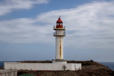 Beyaz yüzlü, kırmızı bluzlu, geleneksel tarzda bir deniz feneri. Yoğun bulutlu mavi gökyüzü. Atlantik Okyanusu kıyısında. Taliarte, Las Palmas, Gran Canaria, Kanarya Adaları, İspanya.