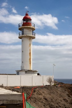 Beyaz yüzlü, kırmızı bluzlu, geleneksel tarzda bir deniz feneri. Yoğun bulutlu mavi gökyüzü. Atlantik Okyanusu kıyısında. Taliarte, Las Palmas, Gran Canaria, Kanarya Adaları, İspanya.