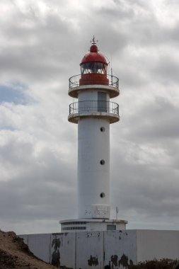 Beyaz yüzlü, kırmızı bluzlu, geleneksel tarzda bir deniz feneri. Yoğun bulutlu mavi gökyüzü. Atlantik Okyanusu kıyısında. Taliarte, Las Palmas, Gran Canaria, Kanarya Adaları, İspanya.