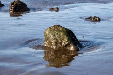 Sahildeki Atlantik okyanus suyu, güneş ışığını ve mavi gökyüzünü yansıtıyor. Güneş ışığında birkaç kaya parçası. Telde, Las Palmas, Gran Canaria, Kanarya Adaları, İspanya.