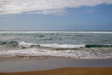 Atlantik Okyanusu 'nun kumlu sahilinde dalgalar ve köpüklerle dolu bir sahil. Kış mevsiminde beyaz bulutlu mavi gökyüzü. Las Palmas de Gran Kanarya, Kanarya Adaları, İspanya.