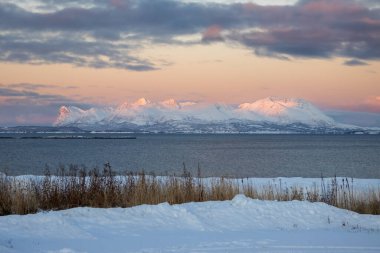 Norveç Denizi kıyısında karla kaplı bir manzara. Arka plandaki dağlar, güneşin doğuşuyla aydınlanıyor. Bulutları olan renkli bir gökyüzü. Sert Stad, Norveç.