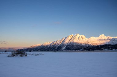 Arka planda doğa ve yüksek dağlar, sonbaharın sonlarında her şey karla kaplıydı. Güneş doğarken renkli kar yağar. Mavi gökyüzü. Hamnvik, Norveç.