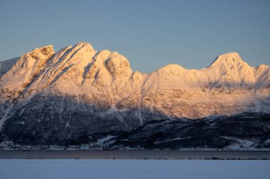 Arka planda doğa ve yüksek dağlar, sonbaharın sonlarında her şey karla kaplıydı. Güneş doğarken renkli kar yağar. Mavi gökyüzü. Hamnvik, Norveç.