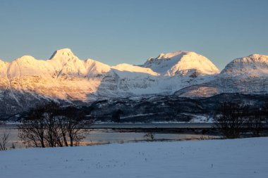 Arka planda doğa ve yüksek dağlar, sonbaharın sonlarında her şey karla kaplıydı. Norveç denizinin sakin suları. Güneş doğarken gökyüzü rengarenk. Mavi gökyüzü. Hamnvik, Norveç.