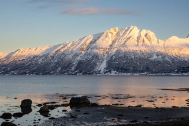 Arka planda doğa ve yüksek dağlar, sonbaharın sonlarında her şey karla kaplıydı. Norveç denizinin sakin suları. Güneş doğarken gökyüzü rengarenk. Mavi gökyüzü. Hamnvik, Norveç.