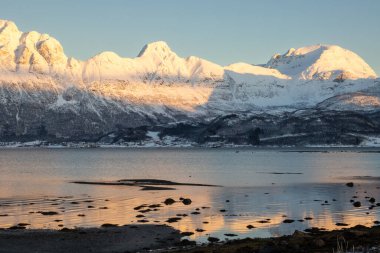 Arka planda doğa ve yüksek dağlar, sonbaharın sonlarında her şey karla kaplıydı. Norveç denizinin sakin suları. Güneş doğarken gökyüzü rengarenk. Mavi gökyüzü. Hamnvik, Norveç.