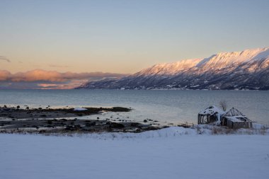 Arka planda doğa ve yüksek dağlar, sonbaharın sonlarında her şey karla kaplıydı. Norveç denizinin sakin suları. Güneş doğarken gökyüzü rengarenk. Mavi gökyüzü. Hamnvik, Norveç.