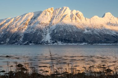 Arka planda doğa ve yüksek dağlar, sonbaharın sonlarında her şey karla kaplıydı. Norveç denizinin sakin suları. Güneş doğarken gökyüzü rengarenk. Mavi gökyüzü. Hamnvik, Norveç.