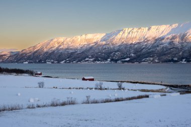 Arka planda doğa ve yüksek dağlar, sonbaharın sonlarında her şey karla kaplıydı. Norveç denizinin sakin suları. Güneş doğarken gökyüzü rengarenk. Mavi gökyüzü. Hamnvik, Norveç.