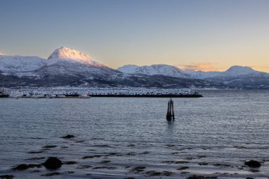 Arka planda doğa ve yüksek dağlar, sonbaharın sonlarında her şey karla kaplıydı. Norveç denizinin sakin suları. Güneş doğarken gökyüzü rengarenk. Mavi gökyüzü. Hamnvik, Norveç.