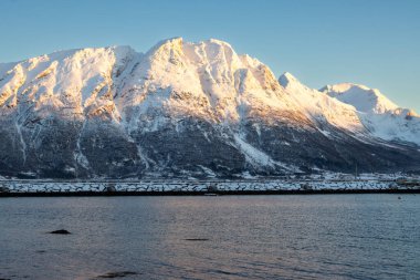 Arka planda doğa ve yüksek dağlar, sonbaharın sonlarında her şey karla kaplıydı. Norveç denizinin sakin suları. Güneş doğarken gökyüzü rengarenk. Mavi gökyüzü. Hamnvik, Norveç.