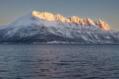 Arka planda doğa ve yüksek dağlar, sonbaharın sonlarında her şey karla kaplıydı. Norveç denizinin sakin suları. Güneş doğarken altın tepeler. Mavi gökyüzü. Hamnvik, Norveç.