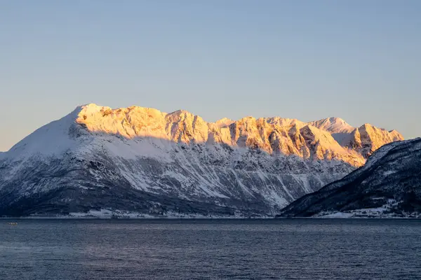 Arka planda doğa ve yüksek dağlar, sonbaharın sonlarında her şey karla kaplıydı. Norveç denizinin sakin suları. Güneş doğarken altın tepeler. Mavi gökyüzü. Hamnvik, Norveç.