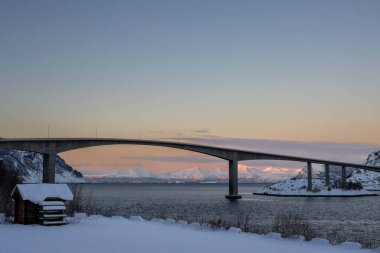Sonbaharın sonlarında karla kaplı bir ülke. Başka bir adaya köprü bağlantısı. Arka plandaki dağlar kutup günbatımının renkleriyle. Krakrohamn, Norveç.