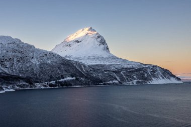 Arka planda doğa ve yüksek dağlar, sonbaharın sonlarında her şey karla kaplıydı. Norveç denizinin sakin suları. Güneş doğarken altın tepeler. Mavi gökyüzü. Hamnvik, Norveç.