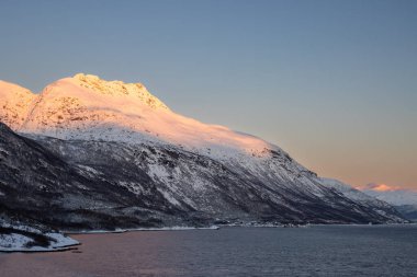 Arka planda doğa ve yüksek dağlar, sonbaharın sonlarında her şey karla kaplıydı. Norveç denizinin sakin suları. Güneş doğarken altın tepeler. Mavi gökyüzü. Hamnvik, Norveç.