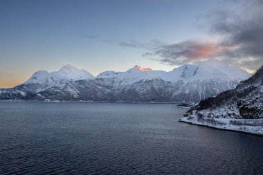 Arka planda doğa ve yüksek dağlar, sonbaharın sonlarında her şey karla kaplıydı. Norveç denizinin sakin suları. Güneş doğarken altın tepeler. Mavi gökyüzü. Hamnvik, Norveç.