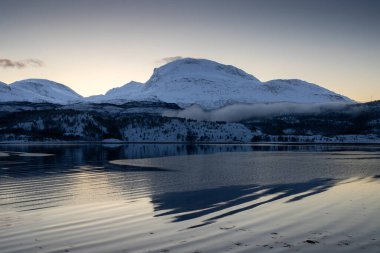Sonbaharın sonlarında kutup günbatımında kar yağan doğa. Norveç denizinin sakin suları. Açık renkli gökyüzü ve bazı açık bulutlar. Loksebotn, Norveç.