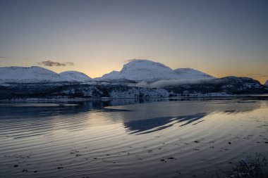 Sonbaharın sonlarında kutup günbatımında kar yağan doğa. Norveç denizinin sakin suları. Açık renkli gökyüzü ve bazı açık bulutlar. Loksebotn, Norveç.
