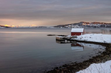 Norveç Körfezi 'nde. Sahildeki kırmızı ahşap ev. Arka plandaki dağlar, Kuzey Kutbu 'nda güneş ışığının dokunduğu yerler. Bulutlu gökyüzü. Harstad, Norveç.