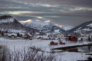 Norveç denizinin körfezi. Sahilde evler ve arka planda dağlar var. Kuzey Kutbu 'nda gün doğumunda güneş ışığının dokunduğu yerler. Bulutlu gökyüzü. Harstad, Norveç.
