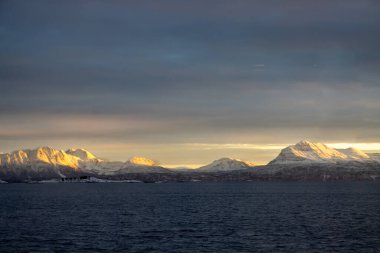 Norveç denizinin sakin suları. Karla kaplı dağlar, kutup gündoğumunda parlak sarı renkler. Renkli gökyüzü. Harstad, Norveç.